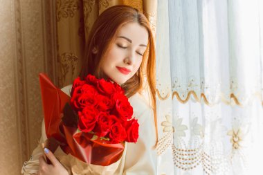 Bej rengi iç dairede bir buket kırmızı gülle romantik bir kadın. Konforlu evde ipek bornozlu bir bayan. Şefkat kavramı ve kadının hissi.