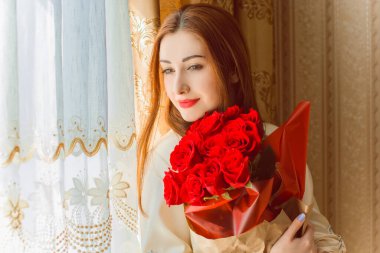 Bej rengi iç dairede bir buket kırmızı gülle romantik bir kadın. Konforlu evde ipek bornozlu bir bayan. Şefkat kavramı ve kadının hissi.