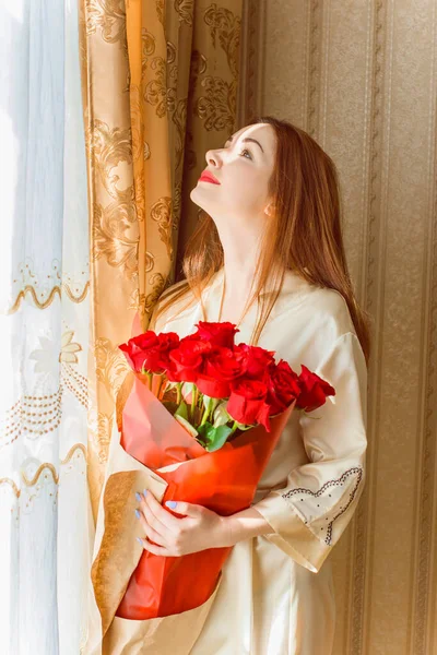 Bej rengi iç dairede bir buket kırmızı gülle romantik bir kadın. Konforlu evde ipek bornozlu bir bayan. Şefkat kavramı ve kadının hissi.