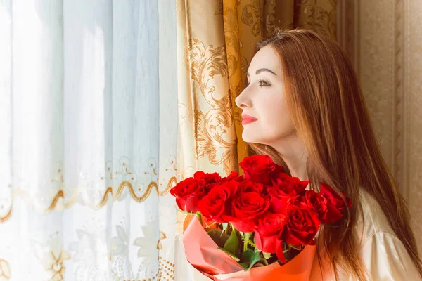 Bej rengi iç dairede bir buket kırmızı gülle romantik bir kadın. Konforlu evde ipek bornozlu bir bayan. Şefkat kavramı ve kadının hissi.