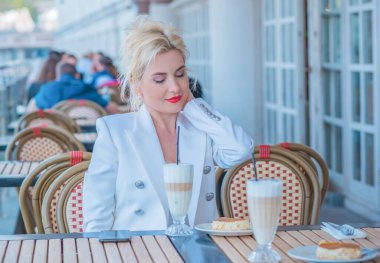 Luxurious woman in white jacket at restaurant. Lady with bright make up and red lips in European cafe drink coffee