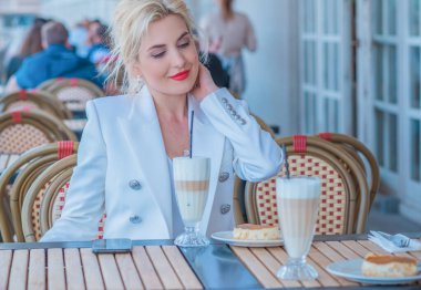 Luxurious woman in white jacket at restaurant. Lady with bright make up and red lips in European cafe drink coffee