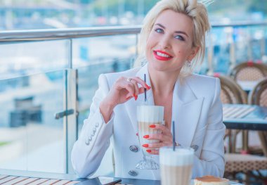 Luxurious woman in white jacket at restaurant. Lady with bright make up and red lips in European cafe drink coffee