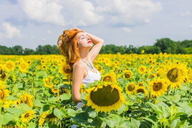 Yazın ortasında tarlanın ortasında, ayçiçekleri içinde beyaz elbiseli, kızıl saçlı, samimi bir kadın. Sağlık konsepti, olumlu duygular ve yaşam tarzı.