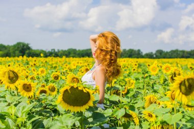 Yazın ortasında tarlanın ortasında, ayçiçekleri içinde beyaz elbiseli, kızıl saçlı, samimi bir kadın. Sağlık konsepti, olumlu duygular ve yaşam tarzı.