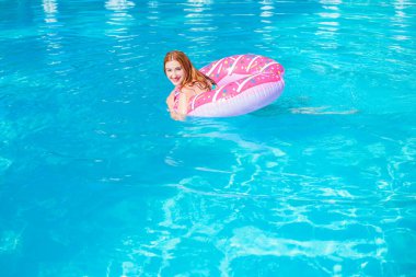 Sıcak ve güneşli bir günde havuzda donut yiyen bir kadın. Yaz tatili cennet gibi. Havuzdaki şişme yatakta bikinili kadın güneşleniyor..