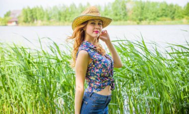 Sazlıkta gülümseyen saman şapkalı olumlu bir genç kadın, klasik tarzda, nehir veya gölde dinleniyor. Amerikan tarzı