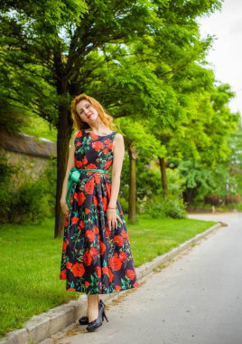 Yazın arka planında gül desenli kırmızı dudaklı, kırmızı elbiseli bir Retro Girl. Bir gül dalının arka planında genç bir kızıl manken. Şık bir kadın. Şık bir yaz elbisesi.