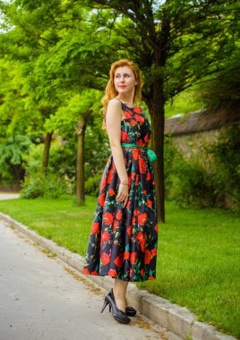 Yazın arka planında gül desenli kırmızı dudaklı, kırmızı elbiseli bir Retro Girl. Bir gül dalının arka planında genç bir kızıl manken. Şık bir kadın. Şık bir yaz elbisesi.