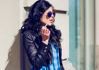 Esmer kadın, mavi gömlekli ve kot pantolonlu. Şehirde sigara içiyor.