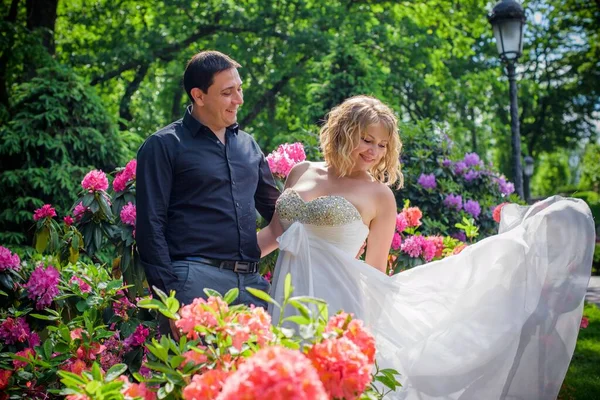美丽的金发碧眼的新娘 在花园里的花朵中 穿着一件长长的浅色连衣裙 形貌华丽 新娘和新郎在散步 — 图库照片