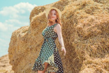 Country tarzı bir karışım, güzel kızıl saçlı bir kadın. Samanlıkta güzel bir bayanın portresi. Yeşil benekli elbiseli ve şapkalı kadın. Moda konsepti