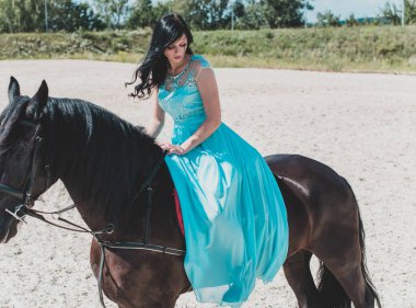 Kırsalda ata binen uzun mavi elbiseli güzel bir genç kadın. Siyah bir at ve kadının portresi. Kırsal bölgede ata binen çekici bir kız.