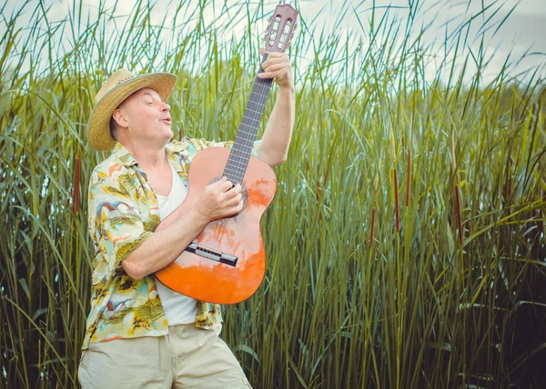 Funny older man in print shirt tourist playing guitar and sing song on nature outdoors, trip to America and pleasure for vacation