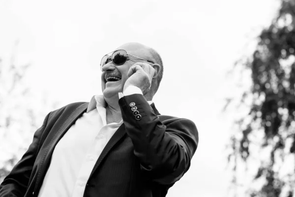 Portrait stylish mature men in white shirt and jacket and blue sunglasses using smart phone. Serving internet via his gadget. Old man using social network technology. Senior man talking on the phone