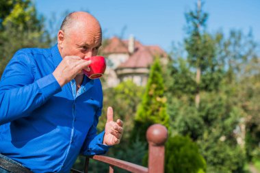 Sabahki kıdemli adam. Elinde bir fincan çay tutarak. Evin bahçesinde kameraya poz veren mavi tişörtlü bir adam. Yaşlılık, içki ve insan konsepti - Mutlu bir çaylı son sınıf öğrencisinin kapanışı