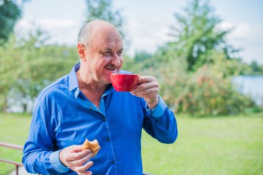 Sabahki kıdemli adam. Elinde bir fincan çay tutarak. Evin bahçesinde kameraya poz veren mavi tişörtlü bir adam. Yaşlılık, içki ve insan konsepti - Mutlu bir çaylı son sınıf öğrencisinin kapanışı