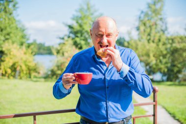 Sabahki kıdemli adam. Elinde bir fincan çay tutarak. Evin bahçesinde kameraya poz veren mavi tişörtlü bir adam. Yaşlılık, içki ve insan konsepti - Mutlu bir çaylı son sınıf öğrencisinin kapanışı