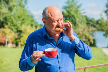 Sabahki kıdemli adam. Elinde bir fincan çay tutarak. Evin bahçesinde kameraya poz veren mavi tişörtlü bir adam. Yaşlılık, içki ve insan konsepti - Mutlu bir çaylı son sınıf öğrencisinin kapanışı