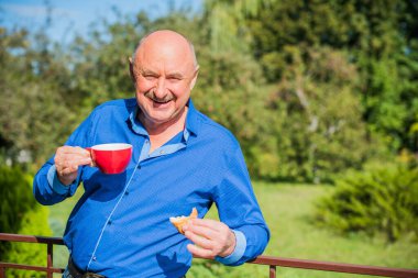 Sabahki kıdemli adam. Elinde bir fincan çay tutarak. Evin bahçesinde kameraya poz veren mavi tişörtlü bir adam. Yaşlılık, içki ve insan konsepti - Mutlu bir çaylı son sınıf öğrencisinin kapanışı