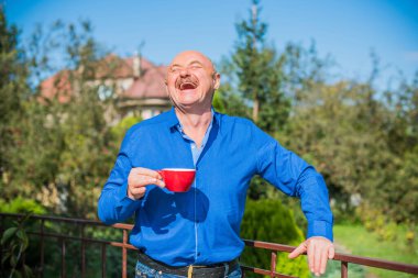 Sabahki kıdemli adam. Elinde bir fincan çay tutarak. Evin bahçesinde kameraya poz veren mavi tişörtlü bir adam. Yaşlılık, içki ve insan konsepti - Mutlu bir çaylı son sınıf öğrencisinin kapanışı