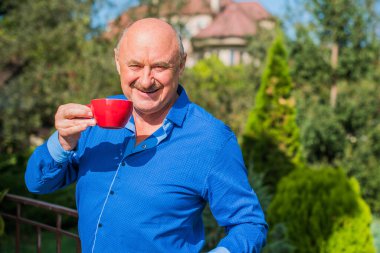 Sabahki kıdemli adam. Elinde bir fincan çay tutarak. Evin bahçesinde kameraya poz veren mavi tişörtlü bir adam. Yaşlılık, içki ve insan konsepti - Mutlu bir çaylı son sınıf öğrencisinin kapanışı