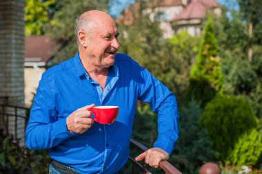 Sabahki kıdemli adam. Elinde bir fincan çay tutarak. Evin bahçesinde kameraya poz veren mavi tişörtlü bir adam. Yaşlılık, içki ve insan konsepti - Mutlu bir çaylı son sınıf öğrencisinin kapanışı