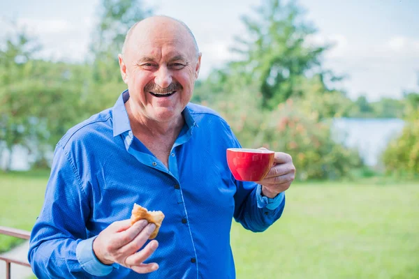 Sabahki kıdemli adam. Elinde bir fincan çay tutarak. Evin bahçesinde kameraya poz veren mavi tişörtlü bir adam. Yaşlılık, içki ve insan konsepti - Mutlu bir çaylı son sınıf öğrencisinin kapanışı