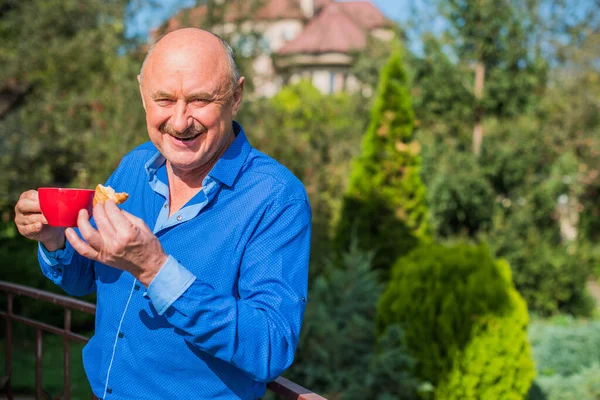 Sabahki kıdemli adam. Elinde bir fincan çay tutarak. Evin bahçesinde kameraya poz veren mavi tişörtlü bir adam. Yaşlılık, içki ve insan konsepti - Mutlu bir çaylı son sınıf öğrencisinin kapanışı
