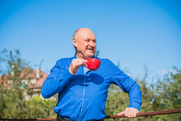 Sabahki kıdemli adam. Elinde bir fincan çay tutarak. Evin bahçesinde kameraya poz veren mavi tişörtlü bir adam. Yaşlılık, içki ve insan konsepti - Mutlu bir çaylı son sınıf öğrencisinin kapanışı