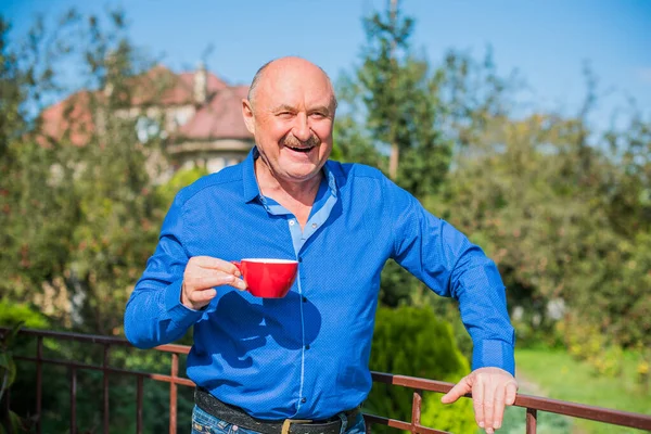 Sabahki kıdemli adam. Elinde bir fincan çay tutarak. Evin bahçesinde kameraya poz veren mavi tişörtlü bir adam. Yaşlılık, içki ve insan konsepti - Mutlu bir çaylı son sınıf öğrencisinin kapanışı
