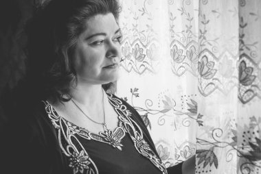 Perdelerin önünde orta yaşlı bir kadın ve pencereden manzaraya bakıyor. Yalnız ve olgun kadınlar, kadınlar arasında sorunlar.