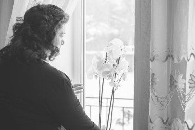 Perdelerin önünde orta yaşlı bir kadın ve pencereden manzaraya bakıyor. Yalnız ve olgun kadınlar, kadınlar arasında sorunlar.