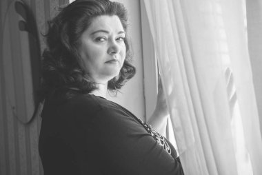 Perdelerin önünde orta yaşlı bir kadın ve pencereden manzaraya bakıyor. Yalnız ve olgun kadınlar, kadınlar arasında sorunlar.