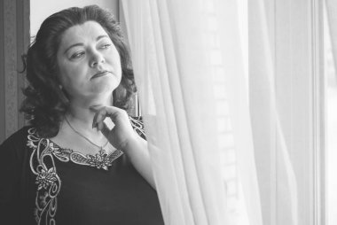 Perdelerin önünde orta yaşlı bir kadın ve pencereden manzaraya bakıyor. Yalnız ve olgun kadınlar, kadınlar arasında sorunlar.