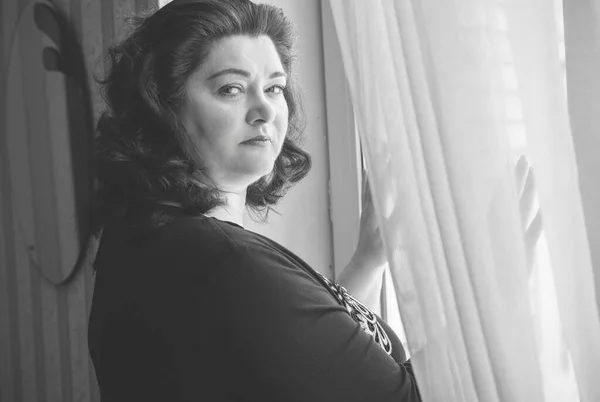 Perdelerin önünde orta yaşlı bir kadın ve pencereden manzaraya bakıyor. Yalnız ve olgun kadınlar, kadınlar arasında sorunlar.