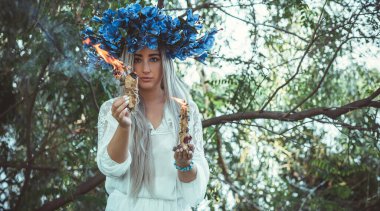 Beautiful girl in wreath of flowers in forest. Portrait of Young beautiful woman wearing white bride dress. Young pagan Slavic girl conduct ceremony on Midsummer. Earth Day