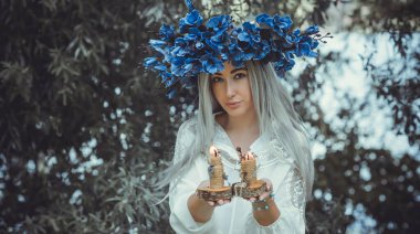 Beautiful girl in wreath of flowers in forest. Portrait of Young beautiful woman wearing white bride dress. Young pagan Slavic girl conduct ceremony on Midsummer. Earth Day
