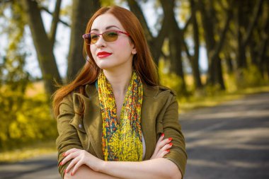 Genç, sevimli, kızıl saçlı bir kadın şık bir şekilde ceket, atkı, yeşil ve kırmızı gözlük takıyor. Bayan sonbahar ağaçlarının korusunda yürüyor.