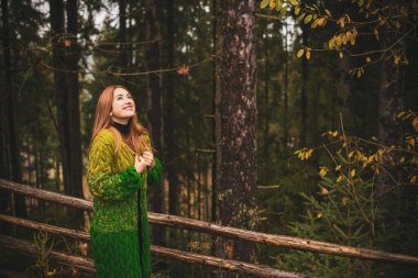 Seyahat konsepti. Kızıl saçlı kadın, soğuk havada dağlarda ve ormanda dinleniyor. Kadın, kalın yün hırka giyiyor, güzel, modaya uygun kıyafetler giyiyor.