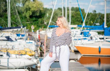 Güzel sarışın kadın, iri yarı, Amerikan ya da Avrupa görünümlü, yatların yanında, hayattan zevk alıyor. Aşırı kilolu, şık bir şekilde çizgili kazak ve beyaz pantolon giymiş genç bir bayan. Doğal güzellik.