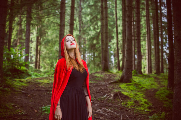 Concept of Halloween. Beautiful and simple costume of little red hood. Mysterious hooded figure in misty forest. Girl in red raincoat. Cosplay Fairy Tale Little Red Riding Hood