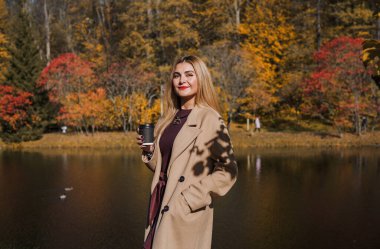 Sonbaharda parkta yürüyen Avrupa ve Ortadoğu görünümlü modern bir kadın. Şık mevsimlik kıyafetler, ılık ışık ve sonbahar renkleri rahat bir yaşam tarzı yaratıyor..