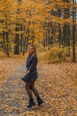 Sonbaharda parkta yürüyen Avrupa ve Ortadoğu görünümlü modern bir kadın. Şık mevsimlik kıyafetler, ılık ışık ve sonbahar renkleri rahat bir yaşam tarzı yaratıyor..