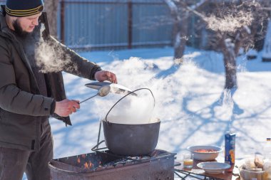 Kışın açık havada sıcak yemek hazırlayan kişi, ateş üzerinde metal bir kazan kullanır. Soğuk havada yemek pişiren elleri yakından izleyin..