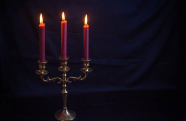 Zarif antika bronz mum tutacağı ve koyu mavi arka planda yumuşak bir şekilde parlayan üç tane burgundy mumu. Metalin dokusunu ve detaylarını vurgulayan klasik, minimalist kompozisyon