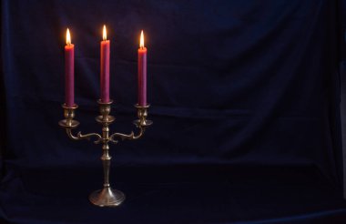 Zarif antika bronz mum tutacağı ve koyu mavi arka planda yumuşak bir şekilde parlayan üç tane burgundy mumu. Metalin dokusunu ve detaylarını vurgulayan klasik, minimalist kompozisyon