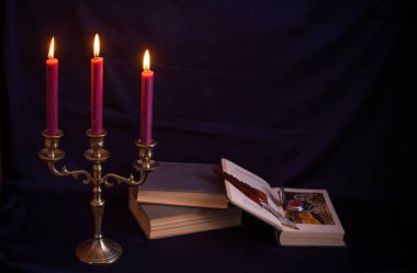 Bronz şamdanlı şarap rengi mumlar, antika kitaplar, tüy kalem ve koyu mavi arka planda eski zarflar.