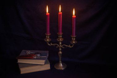 Bronz şamdanlı şarap rengi mumlar, antika kitaplar, tüy kalem ve koyu mavi arka planda eski zarflar.