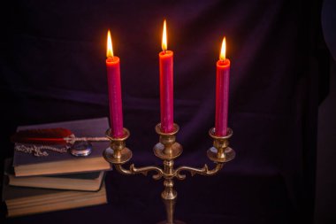 Bronz şamdanlı şarap rengi mumlar, antika kitaplar, tüy kalem ve koyu mavi arka planda eski zarflar.